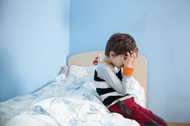 bé trai ôm đầu ngồi bên giường ngủ
